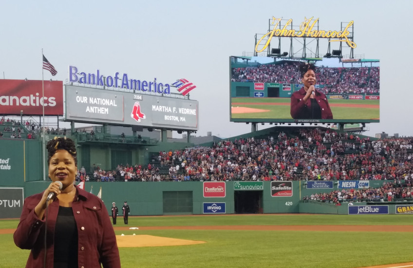 Martha Vedrine singing on the field at Fenway Park, with her image displayed on the jumbotron in the background
