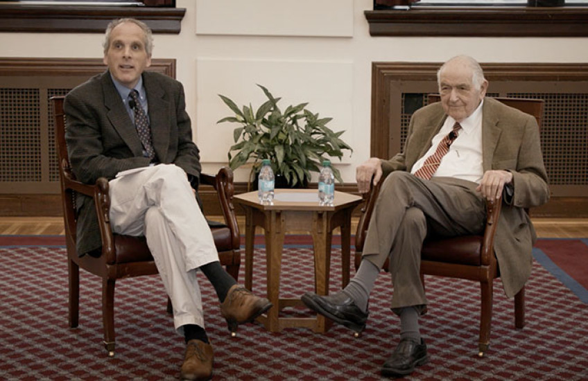 Two men in sports coats and ties sit in armchairs in front of a small table.