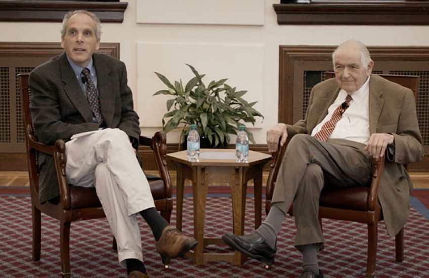 Two men in sports coats and ties sit in armchairs in front of a small table.