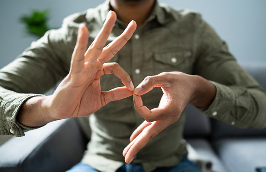 Close-up of two hands belonging to a person of color as they form the ASL sign for “interpreter.” The person’s head is unseen as they sit in a chair in a button-down shirt.