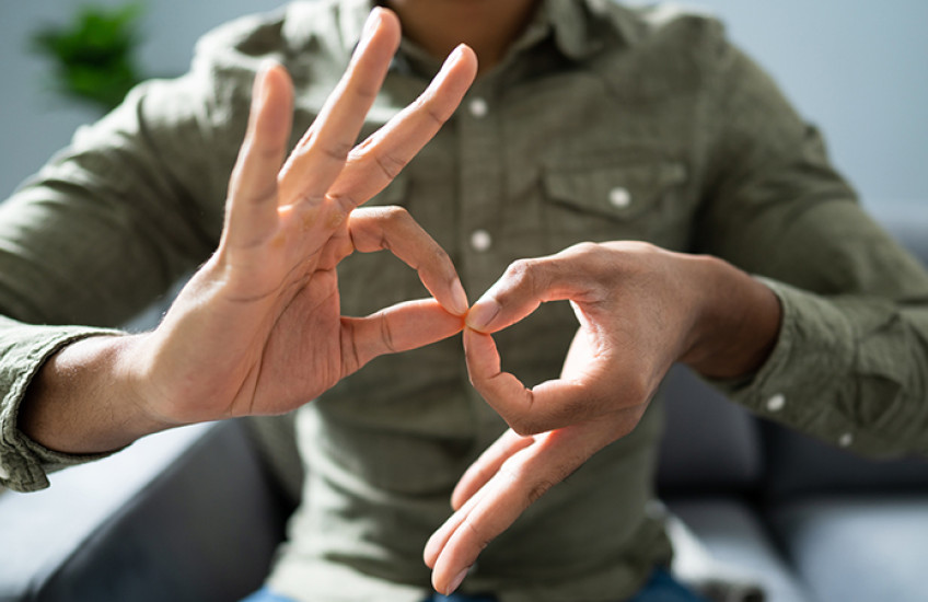 Close-up of two hands belonging to a person of color as they form the ASL sign for “interpreter.” The person’s head is unseen as they sit in a chair in a button-down shirt.