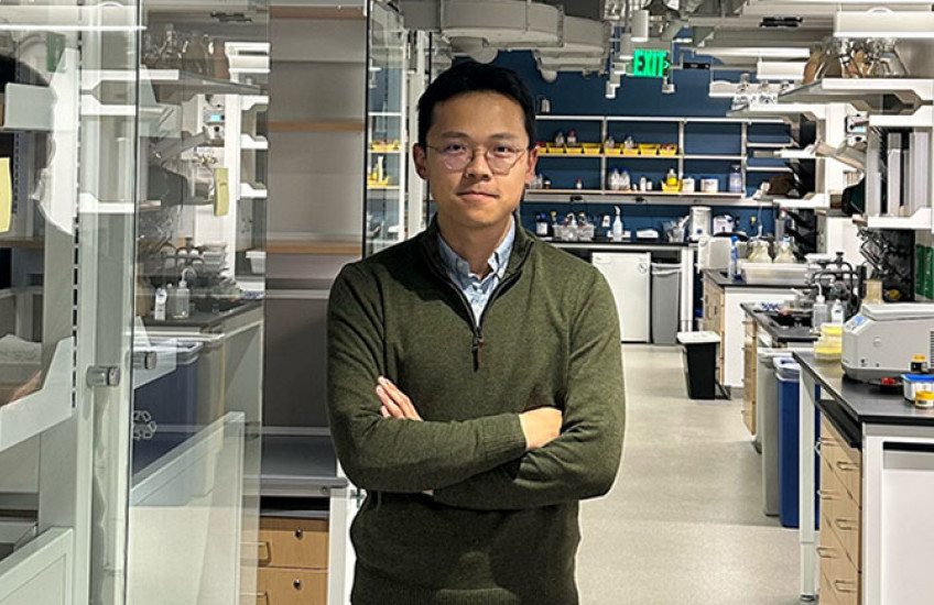 A man stands in a chemistry lab, with machinery on lab benches and reflections on glass partitions. 