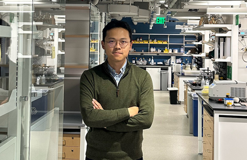 A man stands in a chemistry lab, with machinery on lab benches and reflections on glass partitions.