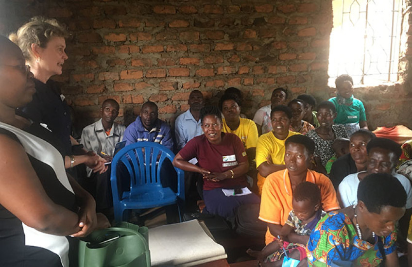 A group of adults seated closely together inside a simple brick-walled room listen attentively as two women stand at the front speaking. The audience includes men and women of different ages, some holding notebooks or bags, creating the feel of a community meeting or training session.