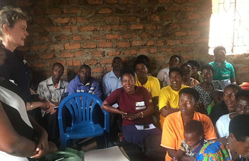 A group of adults seated closely together inside a simple brick-walled room listen attentively as two women stand at the front speaking. The audience includes men and women of different ages, some holding notebooks or bags, creating the feel of a community meeting or training session.