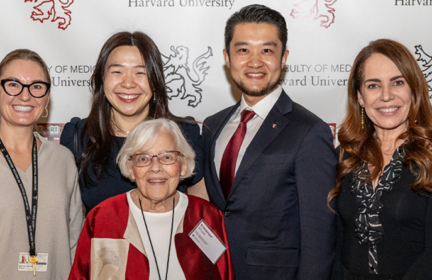 A group of adults stands in front of a backdrop displaying the Harvard University Faculty of Medicine logo. They are smiling and posing for a group photo. The group includes an older woman in a red and beige jacket standing in front. Everyone is dressed in professional or semiformal attire.
