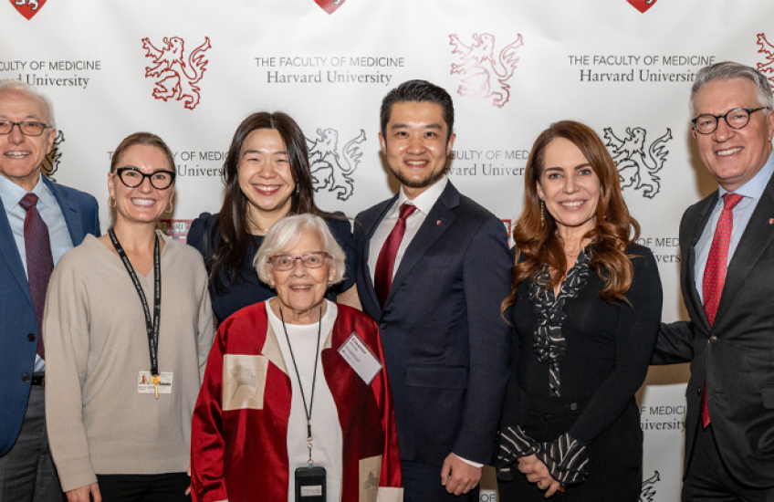 A group of seven adults stands in front of a backdrop displaying the Harvard University Faculty of Medicine logo. They are smiling and posing for a group photo. The group includes an older woman in a red and beige jacket standing in front, with three men and three women standing behind her. Everyone is dressed in professional or semiformal attire, and there are potted plants on either side of the group.