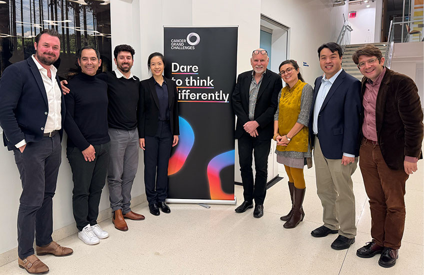 Eight people in business dress pose on either side of a sign that says “Cancer Grand Challenges: Dare to think differently.”
