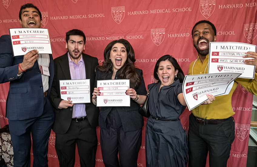 Five young people jump up and down holding their match day signs in front of a Harvard Medical School backdrop.
