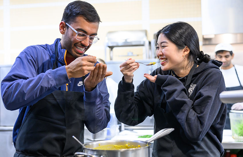 Two young adults wearing aprons taste a spoonful of food from a large pot.