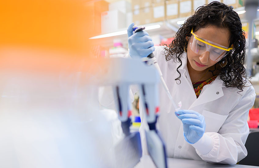 A young woman wearing personal protective equipment (lab coat, safety glasses, gloves) uses a pipette at a lab bench.