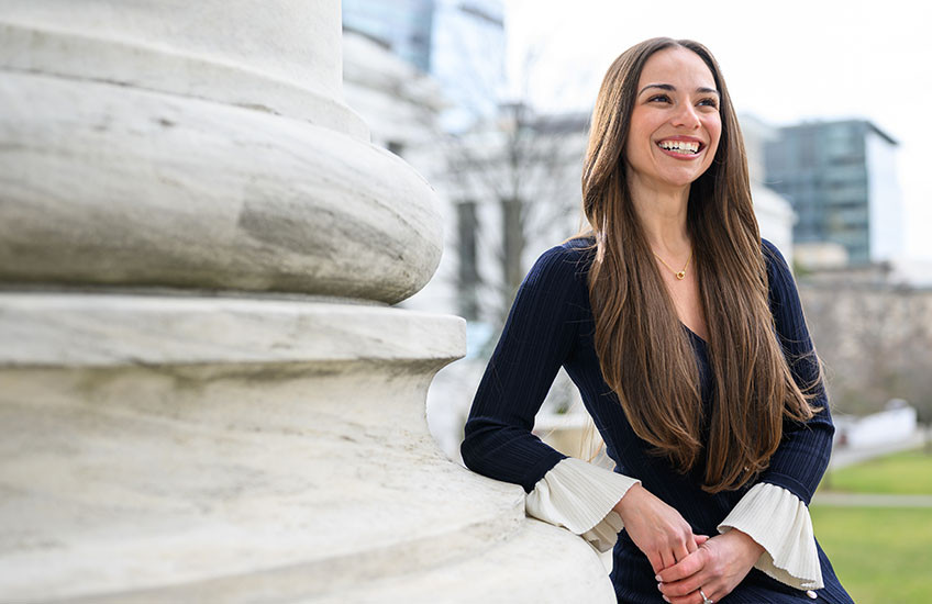 Nikki Pérez smiling, leaning against the base of a column in front of Gordon Hall with the HMS Quad in the background