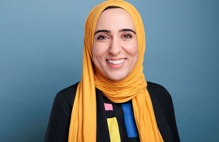 Head and shoulders photo of a woman in a golden yellow hijab and black top with a geometric, colored pattern. 