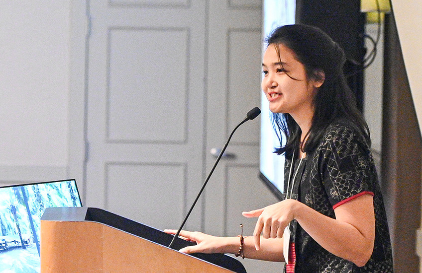 A young woman speaks and gestures at a podium, with four audience members looking on.
