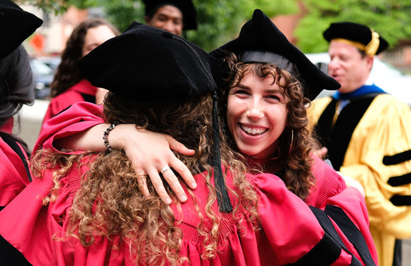 Two women hug and smile in center of photo with other grads and faculty gathered in background.