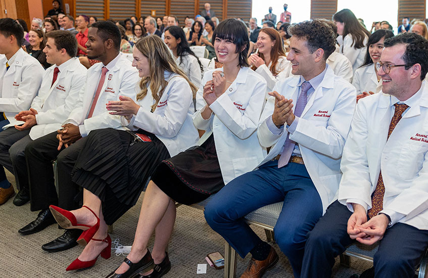 Dozens of students and others sit in rows in a conference room, wearing white coats embroidered with “Harvard Medical School,” smiling and applauding