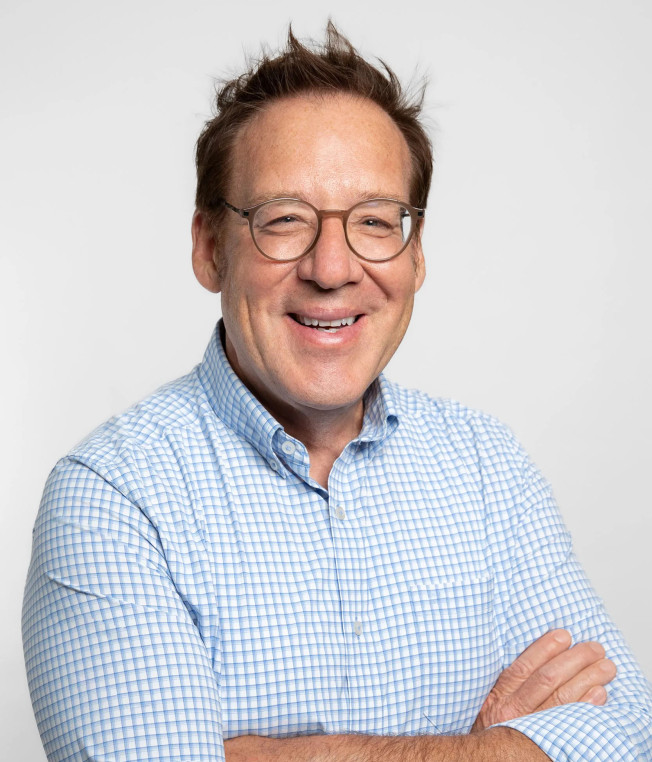 A man with glasses in a blue and white button-down shirt smiles at the camera in front of a gray background.