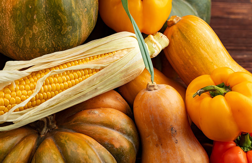A variety of edible squashes on a wooden table along with bell peppers and an open ear of corn.