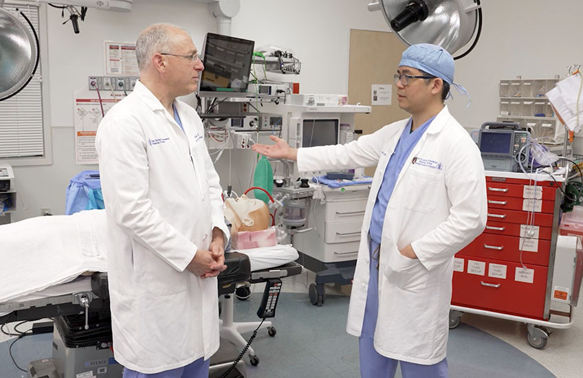 Two men in white coats in a simulated operating room face each other, while a dummy patient lies on an operating table.