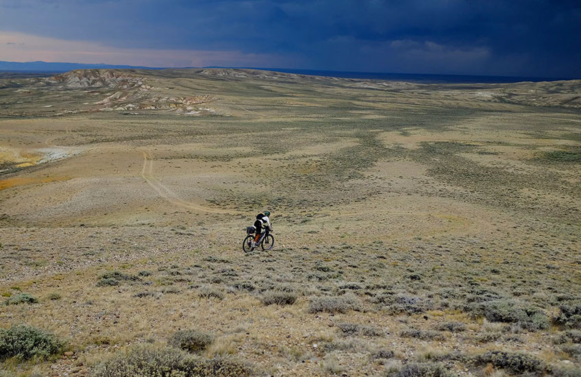 A distant figure on a bicycle traverses wide, open plains under darkening skies.