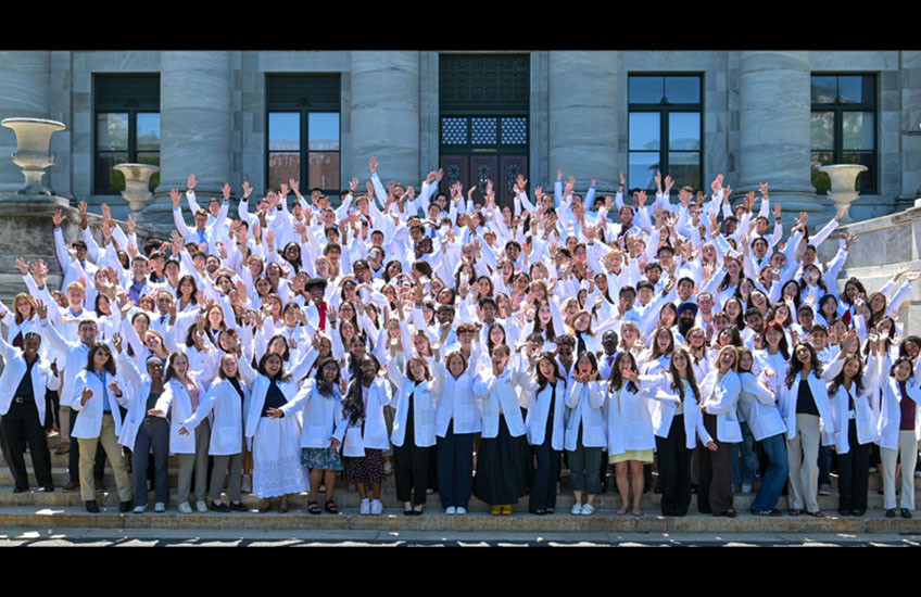 A group of 200 young people stand on the steps of a marble building (Gordon Hall), wearing white coats and cheering.