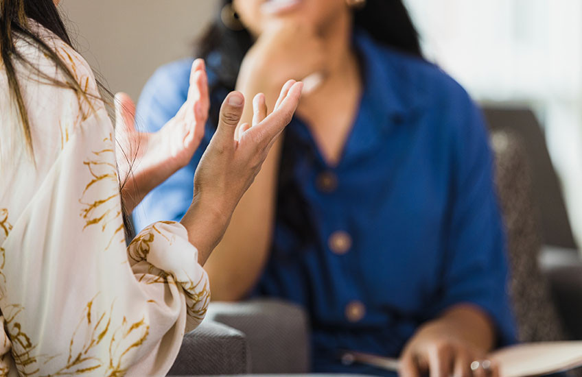 Close-up of the torsos of two people sitting and talking, with one person gesturing with their hands while the other listens with a notebook in their lap.