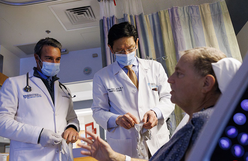 A patient in a hospital bed talks to two surgeons in doctors’ coats and masks.