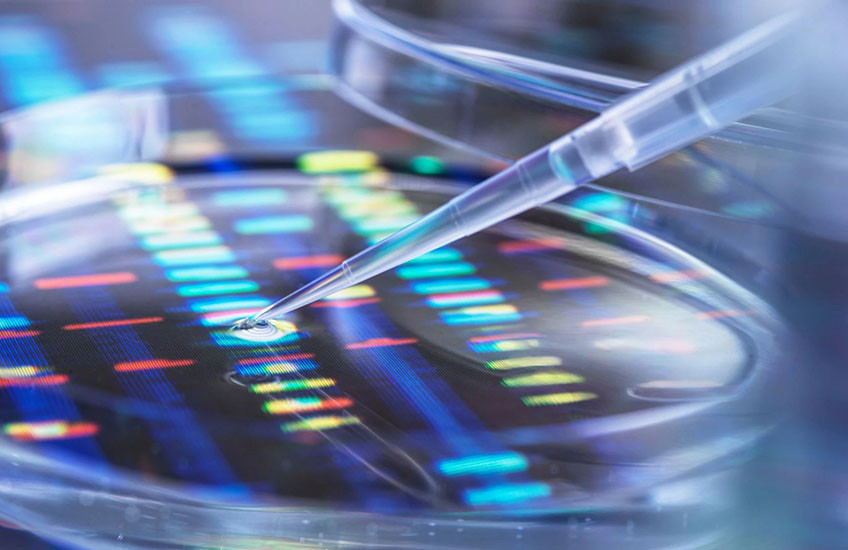 A pipette releases a drop of liquid into a lab dish sitting on a backlit display showing colorful DNA sequencing bands.