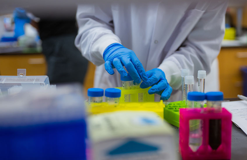 : Scientist wearing a white coat and blue gloves working in a lab setting.
