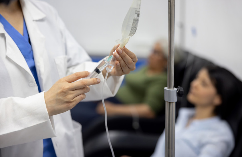 A health care professional administers a medication in a syringe connected to a hanging IV bag beside a seated patient.