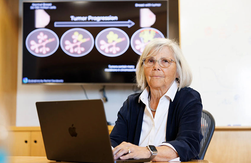 Joan Brugge sits at a desk with a laptop computer in front of a monitor showing a diagram labeled “Tumor Progression.”