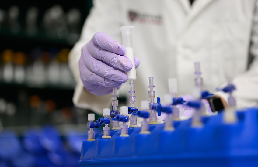 A researcher wearing purple gloves handles a syringe connected to laboratory equipment with multiple valves and tubes arranged in a blue rack