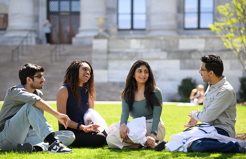 Four young people sit on the grass talking and holding white coats in front of a marble building.