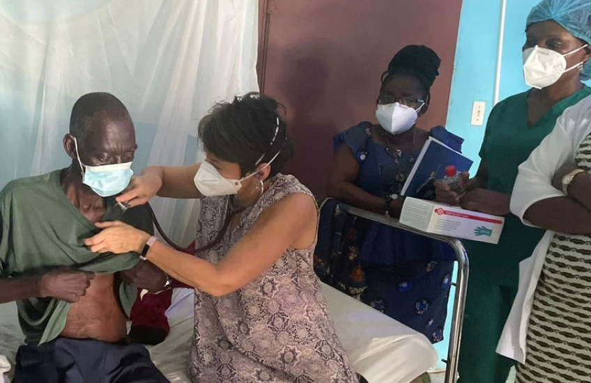 Mukherjee in a print dress wearing a mask holds a stethoscope to the chest of an older man who is wearing a mask while two masked women stand in the background