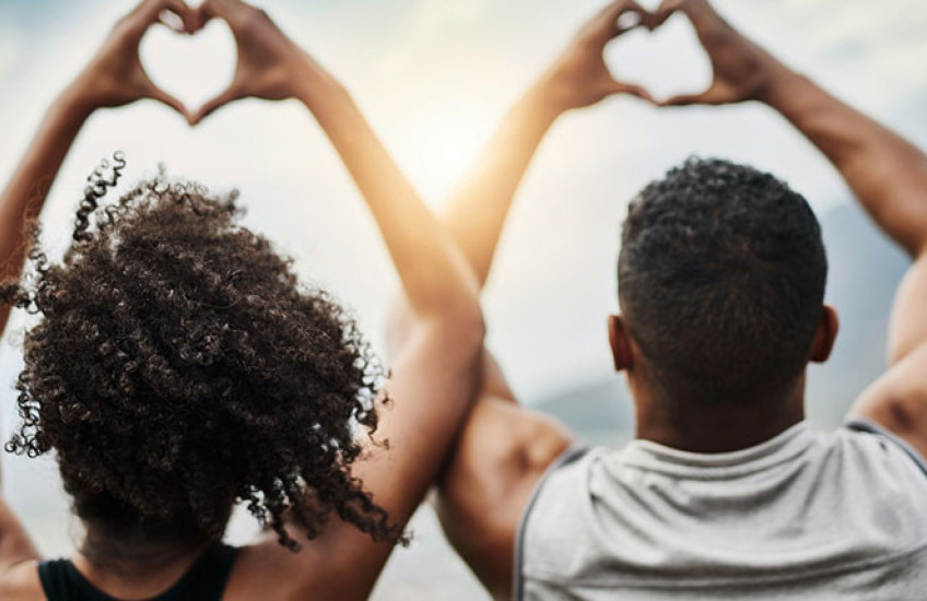 Photo of a Black young man and young woman making heart signs with their hands, backs facing the camera