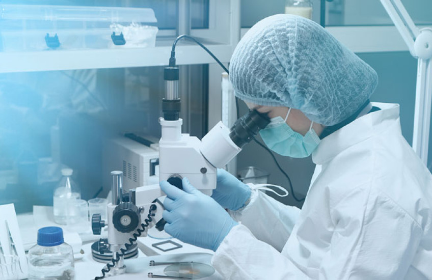 Photo of a female scientist in white lab coat peering into a microscope at a lab bench