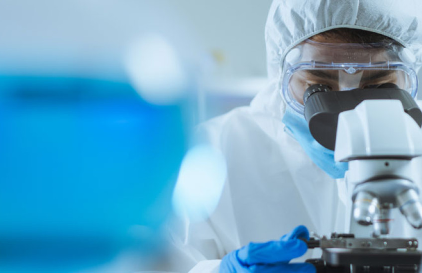 Researcher in white gown and goggles looking into a microscope