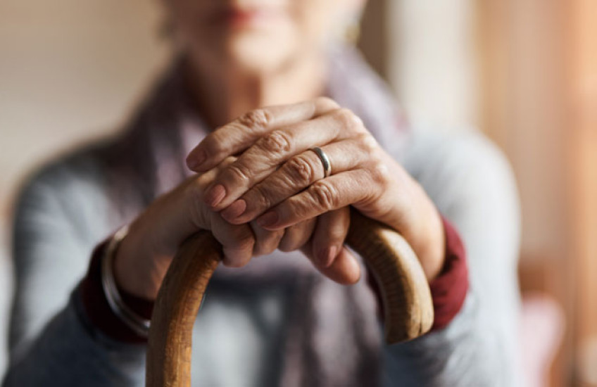 An out-of-focus person sits with their hands on top of a cane in focus in front of them