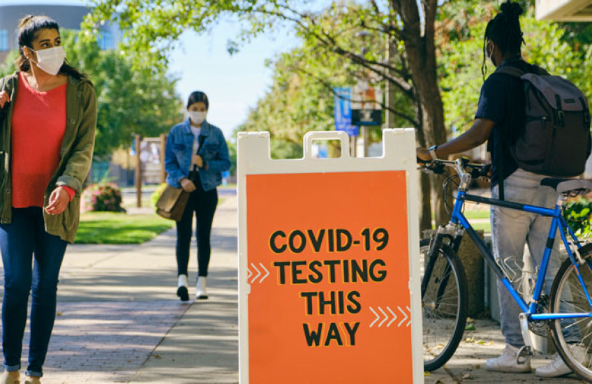 An orange sign that says "COVID-19 testing this way" on a college campus with students walking nearby