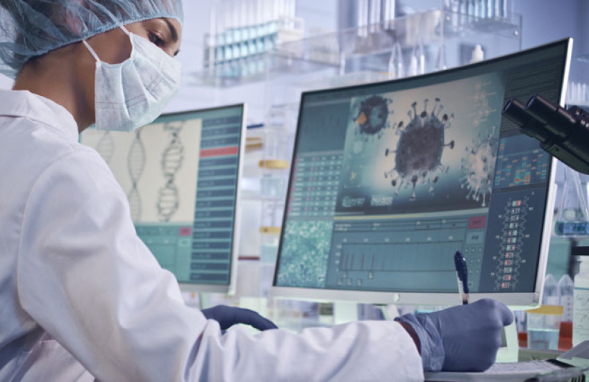 A scientist sits writing notes in front of a computer monitor showing a COVID-19 virus