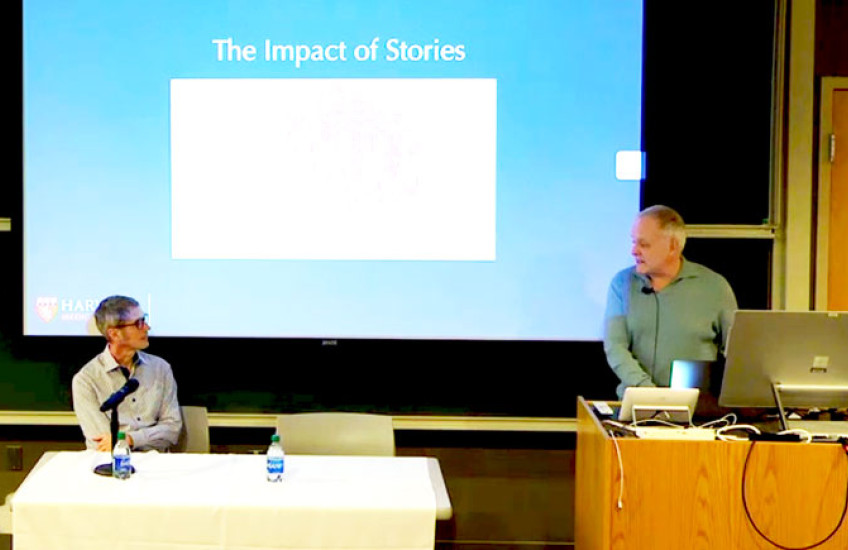 Zoom screen grab of the lecture with Foster seated at a table and Baer speaking from a podium inside a classroom.