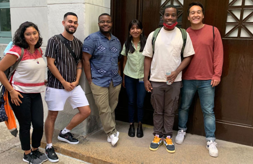 Six students pose for a photo against a stone building and a wooden door