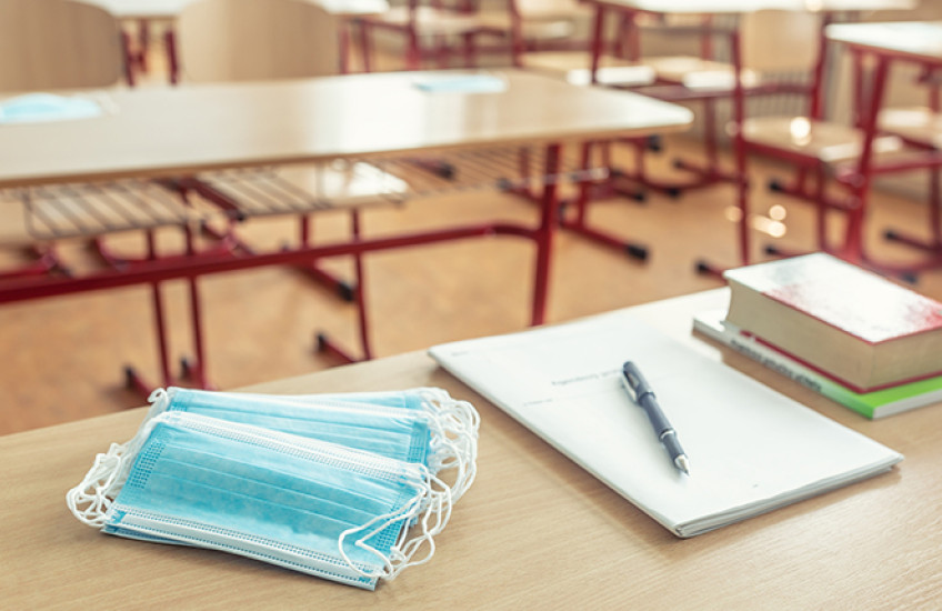 A stack of surgical masks, a pad of paper and pen, and a pile of books sit on a table in an empty classroom