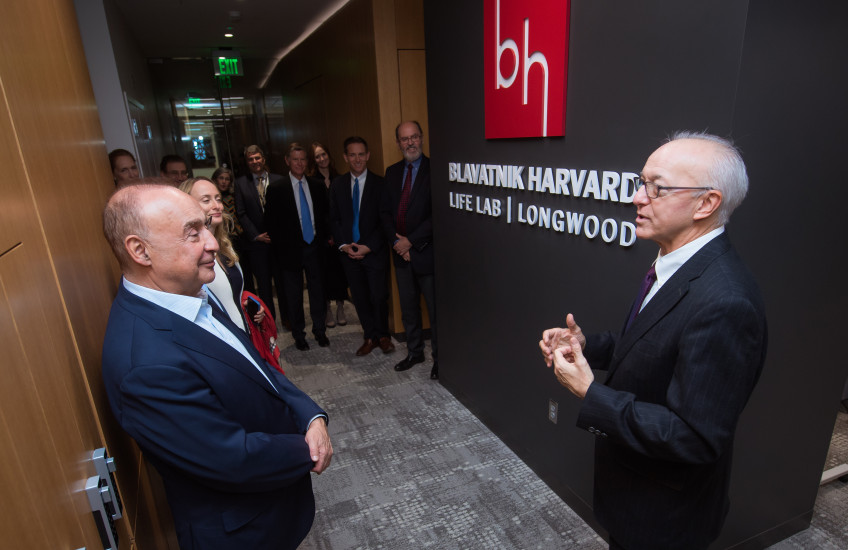 Two men in suits stand in front of a sign with a red square logo with letter B and H above white type on black background that reads Blavatnik Harvard Life Lab Longwood