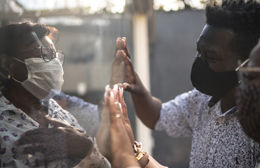 A woman on one side of a window faces a man and a woman on the other side, all three wearing protective masks.