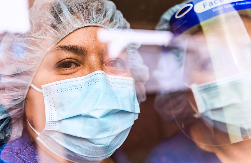 The faces of two people in medical scrubs, protective masks, and head gear seen through a reflection-covered window.