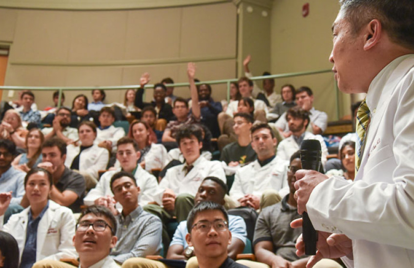 Sideview of Chang in white coat holding mic while speaking to audience of students, many in white coats