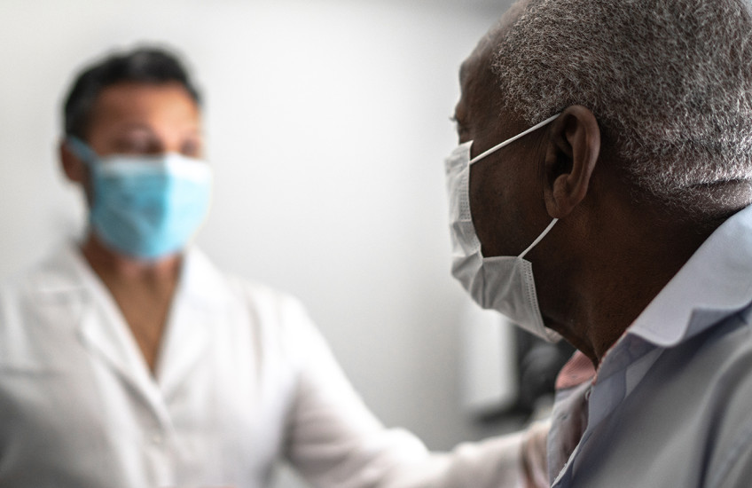 A dark-haired man in a white lab coat sits in a brightly lit room with a gray-haired man in a dress shirt.