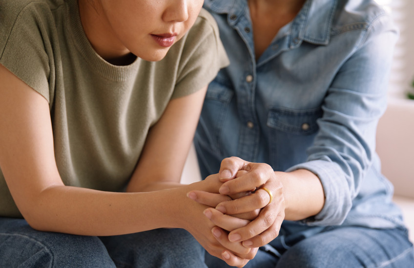 An older person holds a distressed younger person’s hands and shoulder, offering comfort.