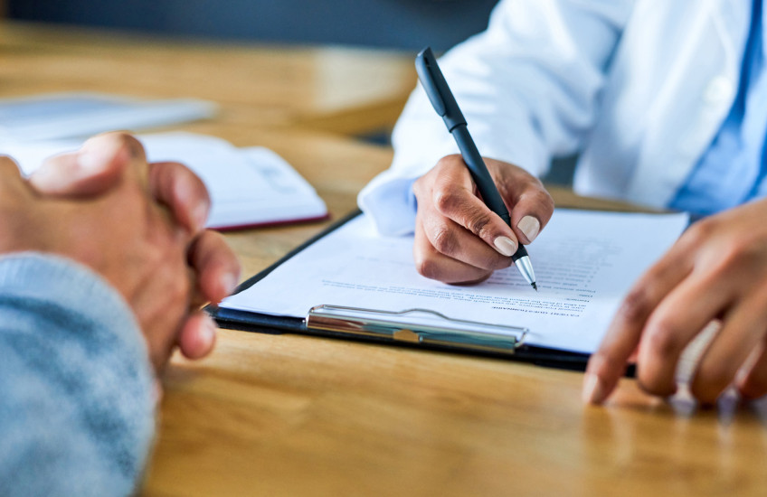 Photograph of doctor's hand writing on a form across from patient with hands folded